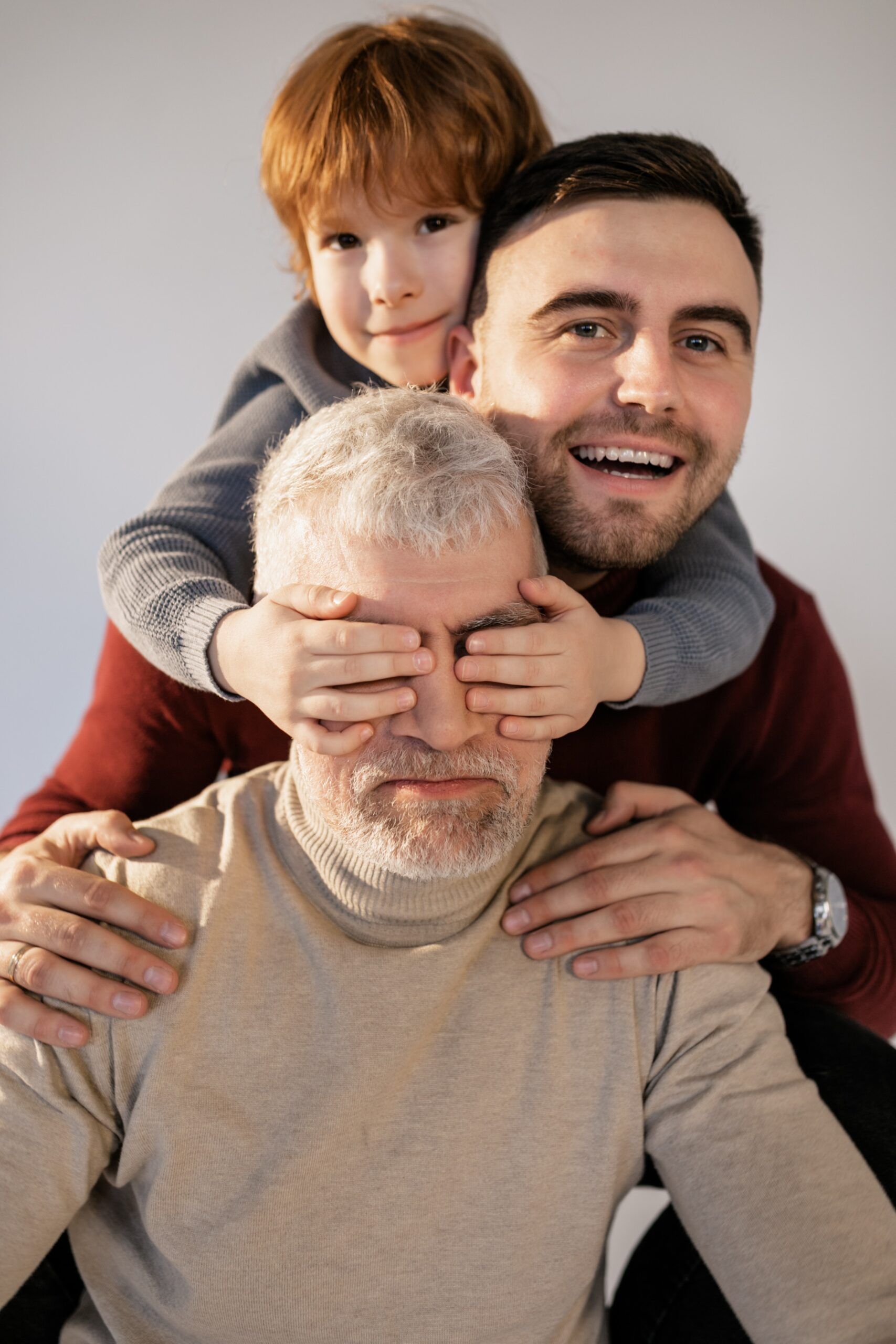 Ein Großvater, Vater und Enkel stehen hintereinander. Der Enkel hält dem Großvater die Augen zu.